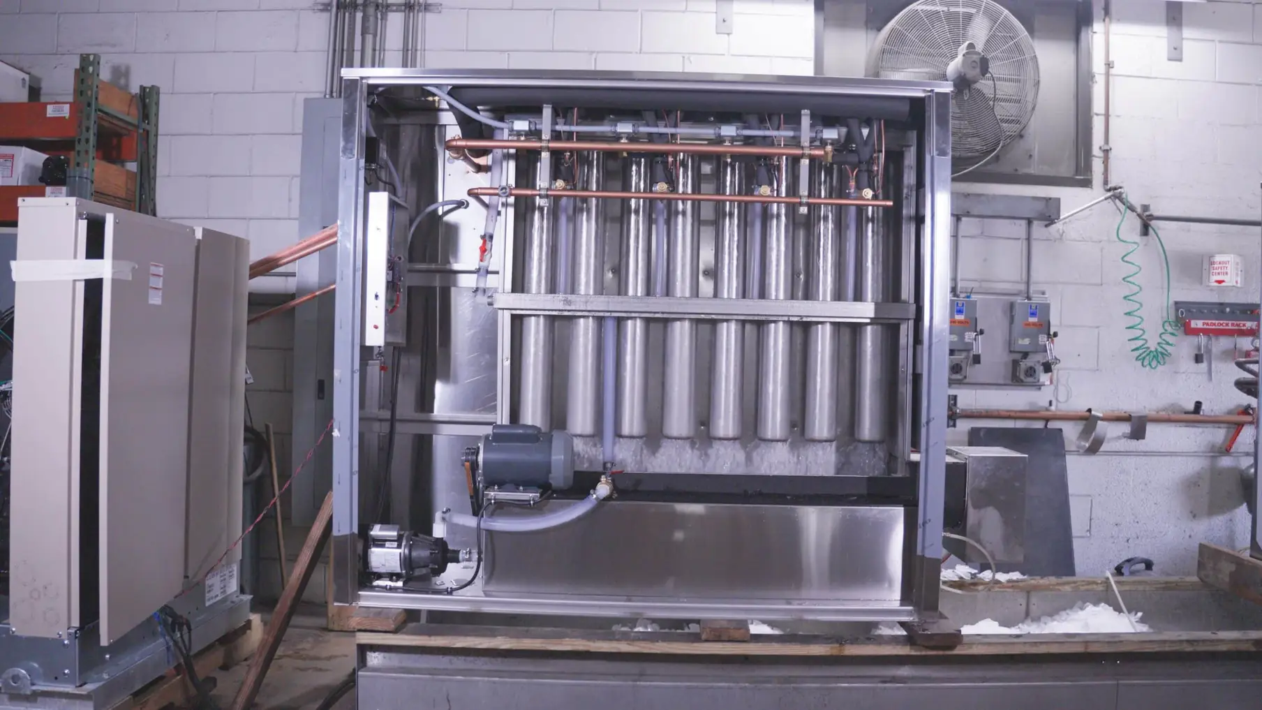 Open ice machine in a brick-walled room showing evaporator tubes and a pump, with a condensing unit at left