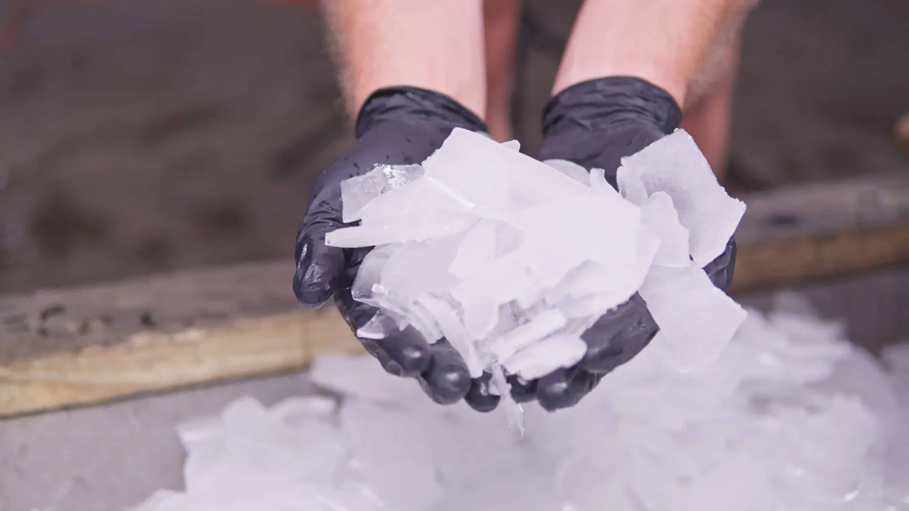 Gloved hands holding a handful of fresh hard cracked ice over a bin full of ice
